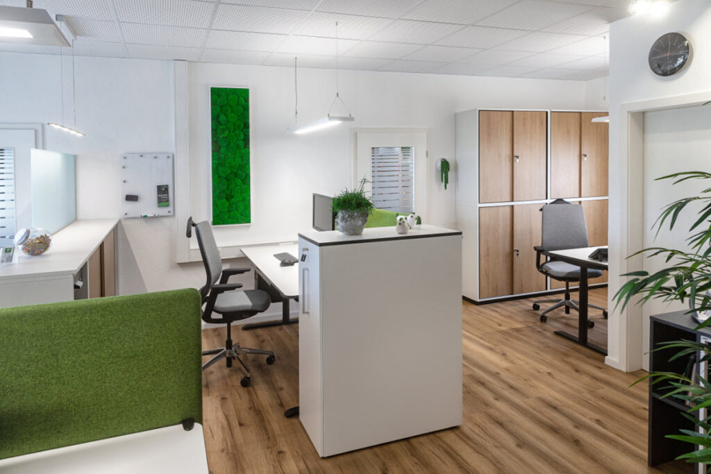 Modern eingerichtetes Büro mit Schreibtischen, Bürostühlen, Holzschränken und grünen Pflanzen an der Wand.