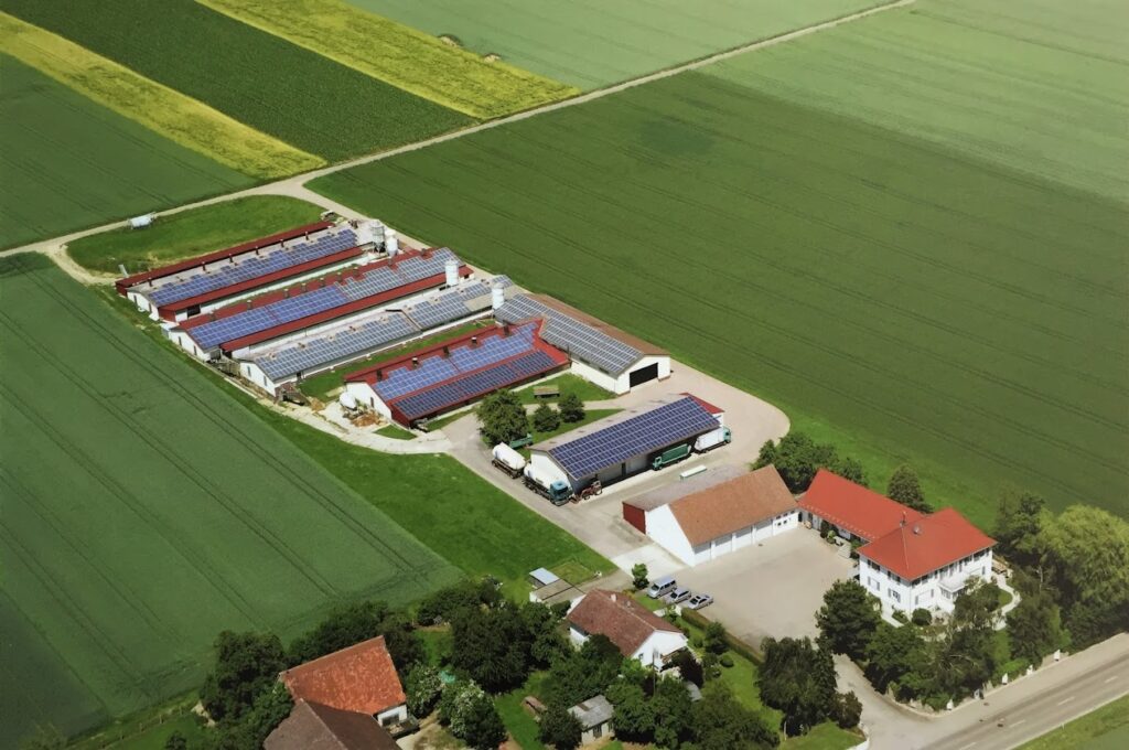 Luftaufnahme des Hühnerhofs Neumair mit mehreren langgestreckten Ställen, die mit Solarpaneelen bedeckt sind, umgeben von grünen Feldern.