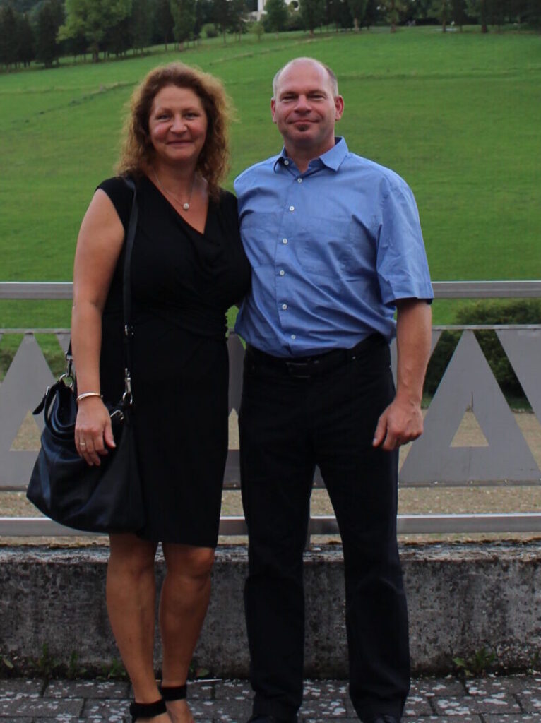 Ein Mann in blauem Hemd und schwarzer Hose steht neben einer Frau in schwarzem Kleid mit schwarzer Handtasche vor einer Brüstung und einer grünen Wiese.